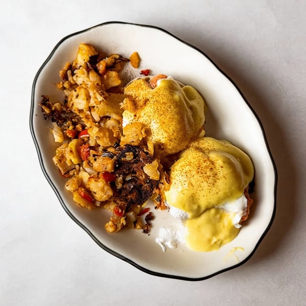 Eggs Benedict with hollandaise sauce and a side of crispy home fries on a white plate.