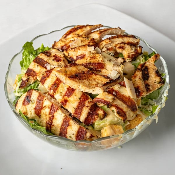 Grilled chicken Caesar salad in a clear glass bowl, with lettuce, croutons, and dressing.