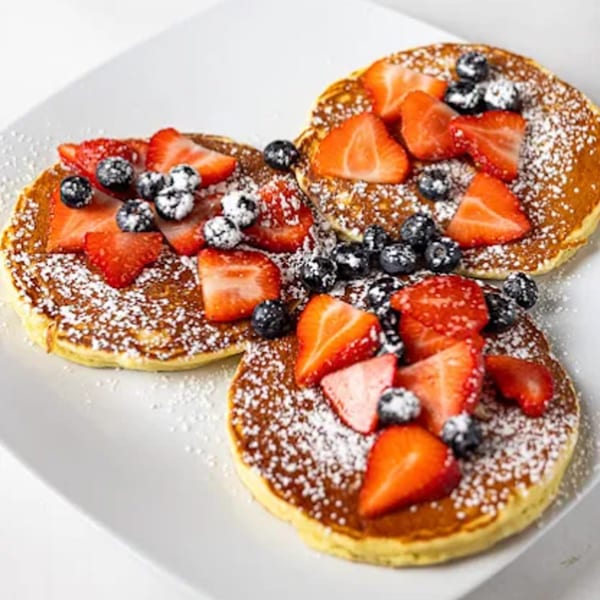 Three Offshore Diner pancakes topped with strawberries, blueberries, and powdered sugar on a white plate.