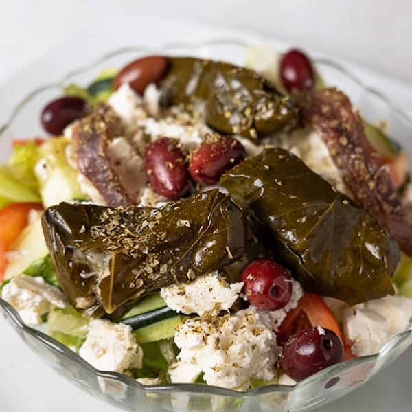 A close-up of a Greek salad with dolmades, feta cheese, olives, and tomatoes in a clear bowl.